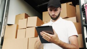 Picture of a delivery man marking off items on his taks list in front of a truck full of boxes.