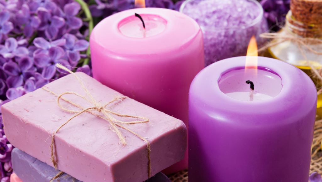 Candle Company Name Ideas - two decorative candles lit, against a background of purple flowers and next to a stack of soaps tied with rustic string